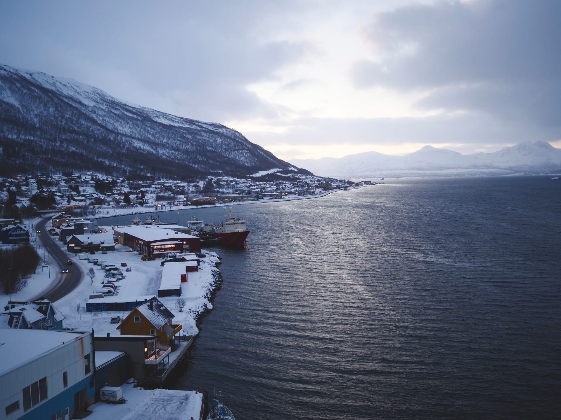 Snødekt kystbygd ved fjorden i vinterhalvlys med eit skip langs kaia