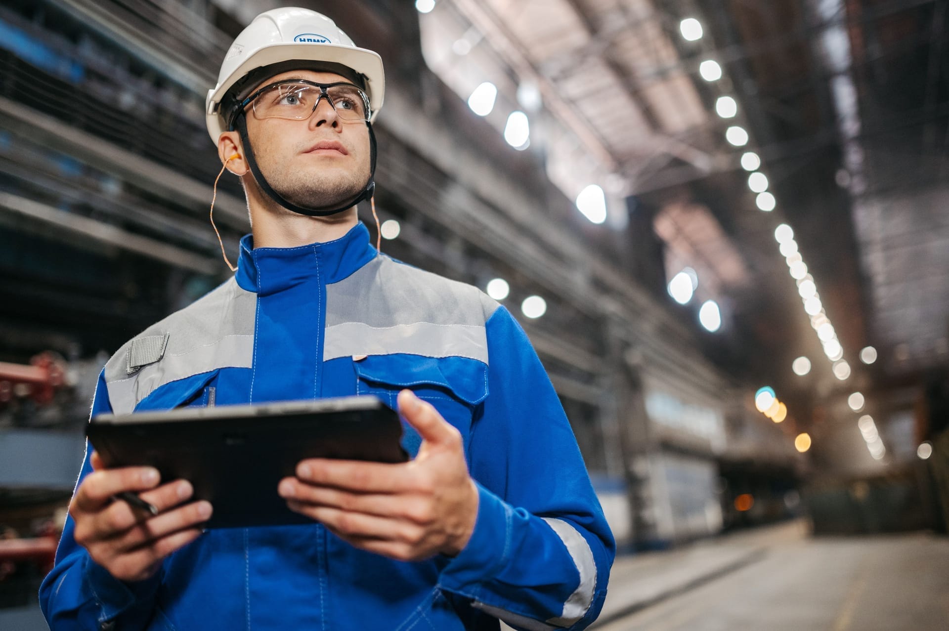 Arbeidar med kvit hjelm og vernebriller held eit nettbrett i ein stor industrihall